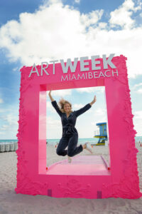 A woman jumps joyfully inside a bright pink “Art Week Miami Beach” photo frame on the sand, with the ocean and a lifeguard tower visible in the background.