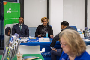 Two CareerSource South Florida representatives sit behind a blue-covered table speaking with an attendee at the Business-Ready Day event. Informational materials are displayed on the table and a green organizational banner stands behind them.