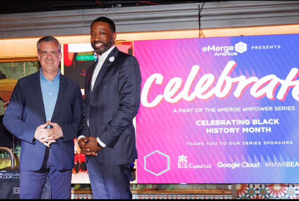 Two men in business attire stand in front of a large digital screen displaying “eMerge Americas Presents Celebrar, Celebrating Black History Month,” posing for a photo at the event.