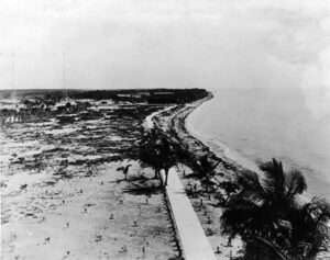 Black-and-white historic aerial view of early Miami Beach around 1910, showing a narrow shoreline, scattered palm trees, a sandy landscape, and a simple wooden walkway running parallel to the ocean.