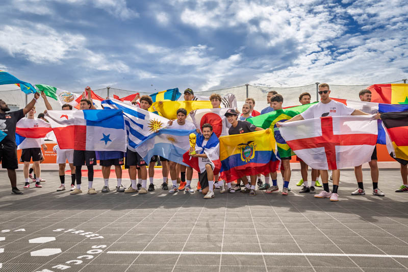 group of 15-20 people holding flags from various countries, man in the middle is kneeling and holding trophy