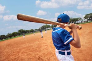 photo taken from behind boy at bat on baseball field, pitcher and opposing team can be seen in front of him