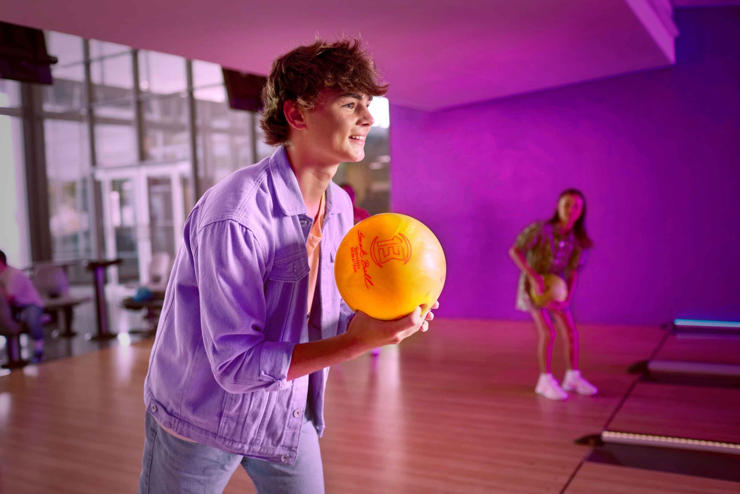 boy holding bowling ball preparing to bowl, a girl holds a bowling ball at another lane in the background