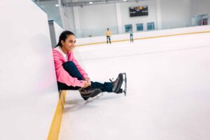 girl putting on ice skates, boy skating in the background on ice rink