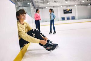boy sitting putting on skates, girls stand on ice rink in the background